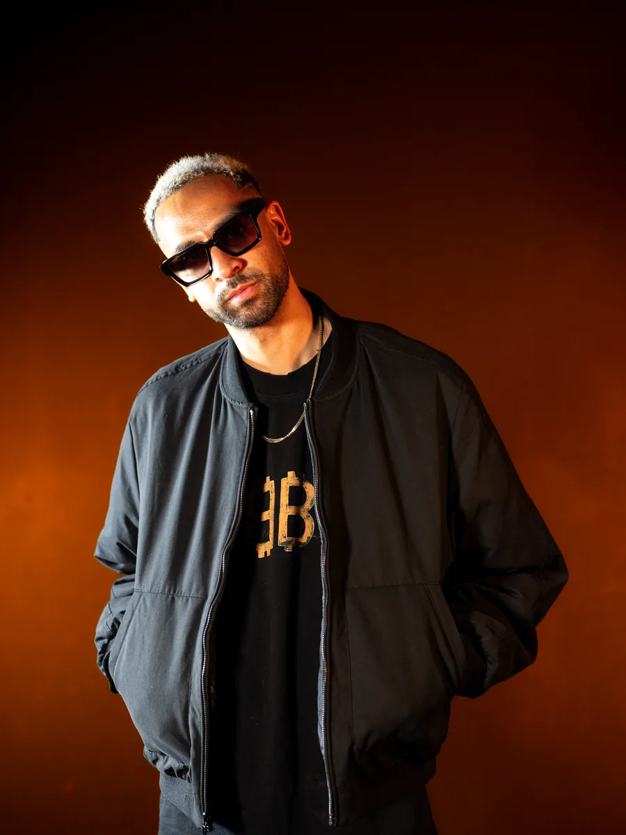 MOKABY in a black bomber jacket and sunglasses against warm amber backlight